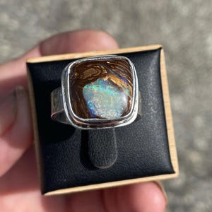 May include: A square-cut opal ring with a silver band. The opal displays a mix of brown, blue, and green hues. The ring is presented on a black display box.