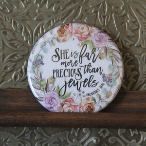 May include: A white round button with a floral design and the text "She is far more precious than jewels Proverbs 3:10".