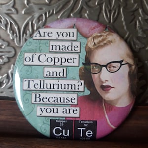 May include: A round button with a turquoise background and a pink border. The button features a vintage-style illustration of a woman with blonde hair wearing glasses. The text on the button reads "Are you made of Copper and Tellurium? Because you are Cu Te".