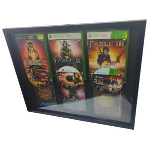 May include: A framed display featuring three Xbox game cases and discs from the Fable series. The cases are green and black, with game titles and character art visible. The discs are displayed below the cases in a black frame.
