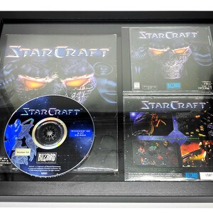 May include: A framed set of StarCraft video game memorabilia, including a CD, a box cover, and two instruction manuals. The box cover features a dark, menacing alien creature with glowing red eyes. The CD is labelled "StarCraft" and features a space-themed design.