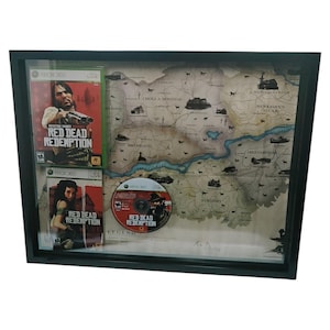 May include: Framed artwork featuring the video game "Red Dead Redemption." The display includes two game cases and a disc, alongside a map of the game's world. The frame is black, and the artwork is set against a neutral background.