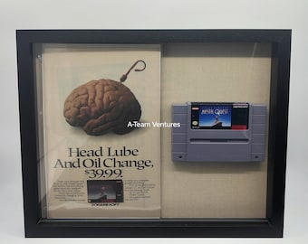 May include: A framed display featuring a vintage video game cartridge for Mystic Quest, a Super Nintendo Entertainment System game. The cartridge is displayed next to a magazine advertisement for the game, featuring a brain with a hook attached to it and the text "Head Lube and Oil Change, $39.99."