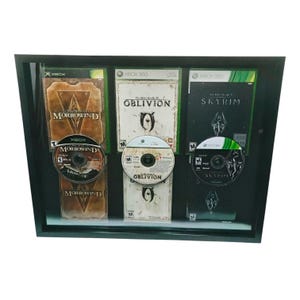 May include: Framed display featuring three Xbox 360 game cases and discs: Morrowind, Oblivion, and Skyrim. The cases are in their original packaging, with game titles and logos visible. The frame is black, and the games are mounted inside.