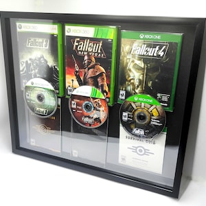 May include: A framed display featuring three Fallout video game cases and discs. The games are for Xbox 360 and Xbox One consoles. The cases are green and black, with game titles visible. The discs are silver and feature game logos.