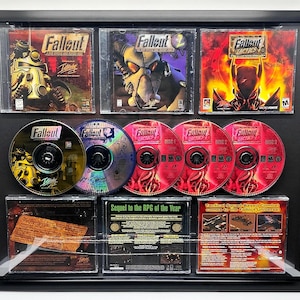 May include: A framed display of the Fallout video game collection. The display includes the game box art for Fallout, Fallout 2, and Fallout Tactics. It also includes the game discs for Fallout Tactics, which are labeled with the game's title and disc number. The display is set against a black background.