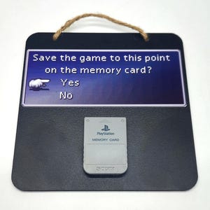 May include: A black chalkboard sign with a white border and a blue and white image of a PlayStation game save screen. The text on the screen reads "Save the game to this point on the memory card? Yes No". Below the image is a gray PlayStation memory card.