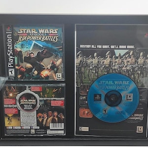 May include: A black framed display case with two PlayStation game cases for Star Wars Episode I: Jedi Power Battles. The game cases are displayed next to a promotional poster for the game. The poster features a blue disc with the game title and a scene of a battle between clone troopers and battle droids. The text on the poster reads "Destroy all you want. We'll make more."