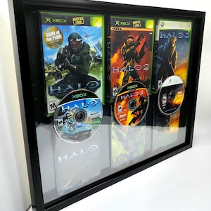 May include: A black framed display case featuring three video game cases and three discs. The cases are for Halo, Halo 2, and Halo 3. The discs are for Halo, Halo 2, and Halo 3. The cases all share a similar design, with a black background and green and orange accents. The discs also have a consistent design, with a black background and green and orange accents.