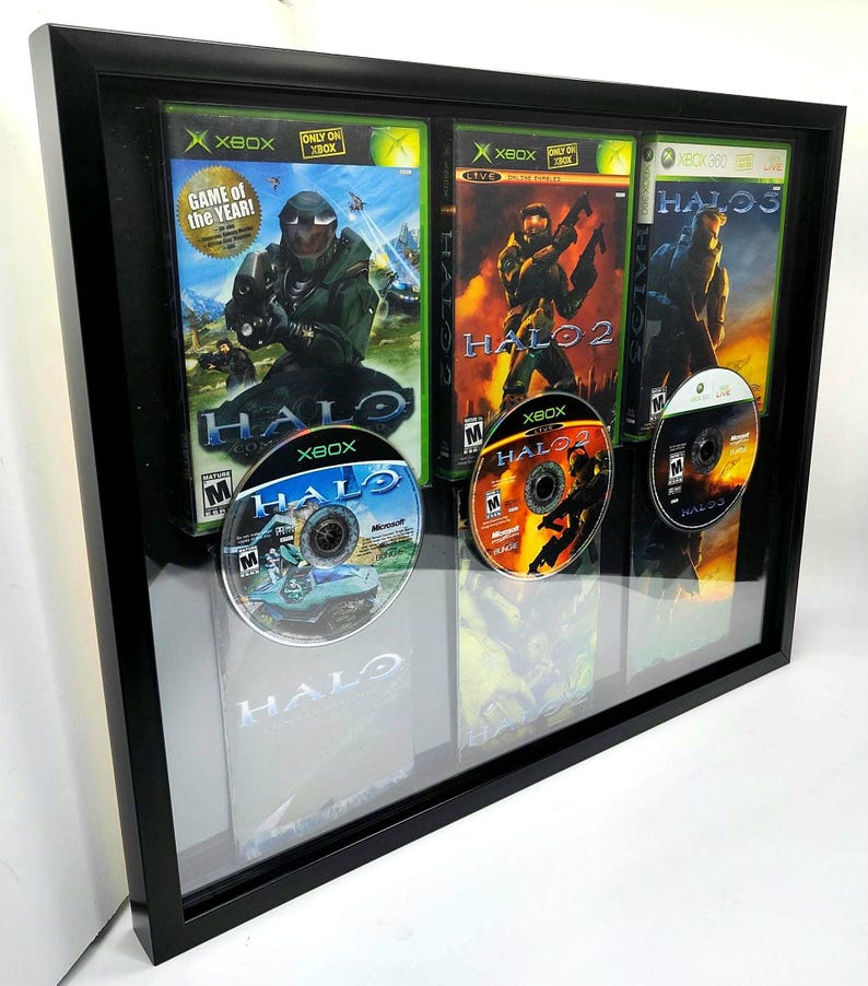 May include: A black framed display case featuring three video game cases and three discs. The cases are for Halo, Halo 2, and Halo 3. The discs are for Halo, Halo 2, and Halo 3. The cases all share a similar design, with a black background and green and orange accents. The discs also have a consistent design, with a black background and green and orange accents.