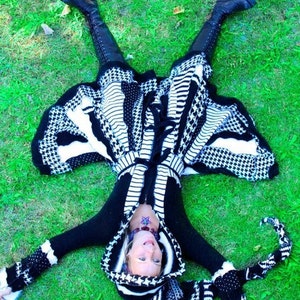 Może przedstawiać: Sweter z kapturem i długimi rękawami w czarno-biały wzór. Sweter jest noszony przez osobę leżącą na plecach na zielonej trawie. Osoba ma na sobie czarne buty i ma tatuaż w kształcie gwiazdy na klatce piersiowej.