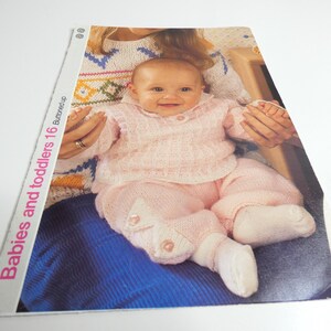 May include: A pink knitted baby outfit with a cardigan and pants. The outfit is being worn by a baby sitting on a person's lap. The text "Babies and toddlers 16 Buttoned up" is visible on the side of the image.