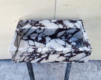 Può includere: Un lavandino in marmo bianco con venature di colore viola scuro. Il lavandino è rettangolare e ha un solo foro di scarico. Il lavandino è montato su un supporto in metallo.