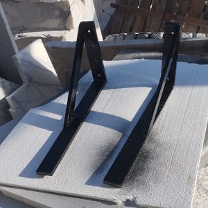 Könnte beinhalten: Zwei schwarze Metall-Regalhalterungen. Jede Halterung hat ein rechtwinkliges Design mit einer flachen Basis und einem vertikalen Stützteil. Die Halterungen werden auf einer weißen Oberfläche mit einem Hintergrund aus Steinplatten gezeigt.