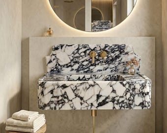 May include: A marble bathroom sink with black and white veining, featuring gold fixtures and a decorative backsplash. A small vase with dried flowers sits on the countertop. A stack of folded beige towels rests on a wooden stool.