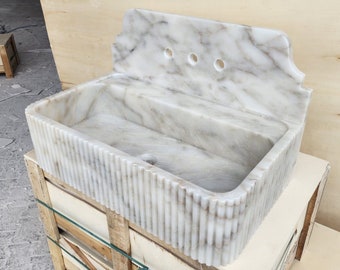 May include: A white marble sink with a fluted edge and a backsplash with three holes. The sink is sitting on a wooden crate.