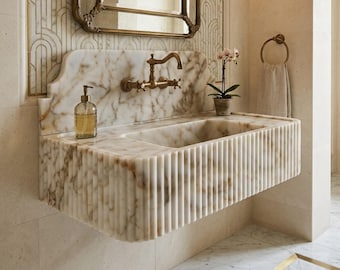 May include: A marble bathroom sink with a fluted front design. The sink has a built-in countertop with a soap dispenser and a gold faucet. A decorative mirror hangs above the sink, and a towel ring is on the right.