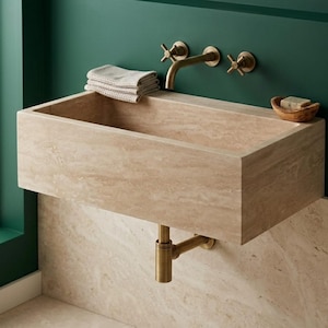 Peut inclure: Un évier rectangulaire en pierre beige avec un robinet et des fixations en laiton. Un petit bol en bois avec du savon est posé sur le côté droit de l'évier. Une serviette beige pliée repose sur le bord de l'évier. L'évier est monté sur un mur beige et vert.