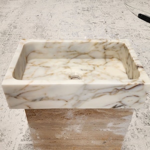 May include: A white marble bathroom sink with gold veining. The sink is rectangular and has a rounded edge. It is sitting on a pedestal made of a light brown stone.