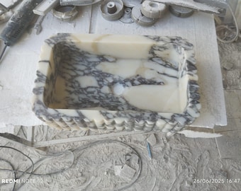 Puede incluir: Un lavabo rectangular de mármol con un patrón veteado crema y gris. El lavabo tiene un borde festoneado y un interior liso. El lavabo es probablemente hecho a mano y es una pieza única para un baño.