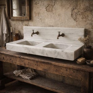 Peut inclure: Un évier double en marbre blanc avec deux robinets en laiton. L'évier est placé contre un mur texturé et repose sur une table en bois rustique. La scène comprend un miroir, une serviette et des objets décoratifs, créant une esthétique de salle de bain vintage.