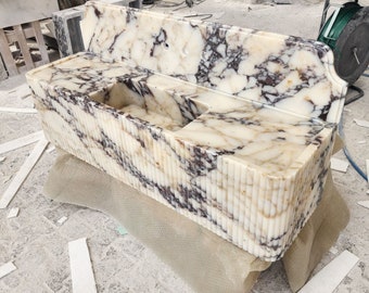 May include: A marble vanity unit with an integrated sink. The vanity is white with dark purple and brown veining. The front features vertical ridges. The sink has a rectangular basin and a small side basin.