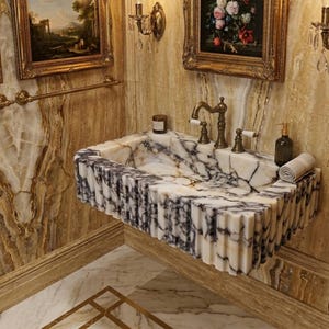 May include: A luxurious marble sink with a fluted design, featuring a mix of white and gray veining. The sink has bronze fixtures and is set against a backdrop of ornate wall paneling and framed artwork. A rolled towel and soap dispenser are visible.
