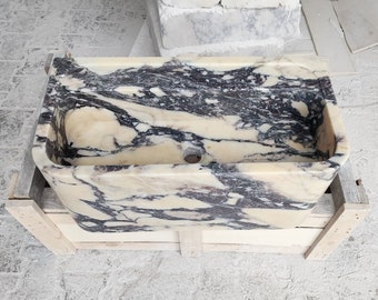 Könnte beinhalten: Ein rechteckiges Marmorwaschbecken mit einem weißen und violetten Marmormuster. Das Waschbecken hat ein Abflussloch in der Mitte und steht auf einer Holzkiste.