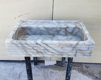 May include: A white marble sink with gold veining, mounted on a black metal stand. The sink has a rectangular shape and a slightly rounded edge.