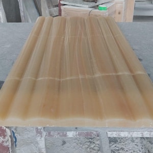 May include: A rectangular, light amber-coloured stone slab with a ribbed, wave-like surface. The stone has a translucent quality, with subtle variations in colour and veining. The slab is likely for architectural or design use.