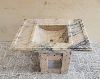 May include: A rectangular marble sink with a light beige and gray veined pattern. The sink has a curved basin and two side ledges. It is set on a rustic wooden stand, showcasing a natural stone design.