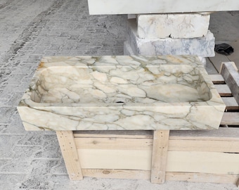 May include: A rectangular sink carved from polished natural stone featuring a cream-colored base with prominent grey and golden-brown veining throughout the surface. The basin has a smooth interior finish and a single centered drain hole at the bottom.