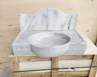 Op de afbeelding: Een witte marmeren wastafel met een ronde kom en een hoge, gebogen achterwand. De steen vertoont over het hele oppervlak natuurlijke grijze aders, met een gladde, gepolijste afwerking op de kom en het vlakke gedeelte rondom de wasbak.