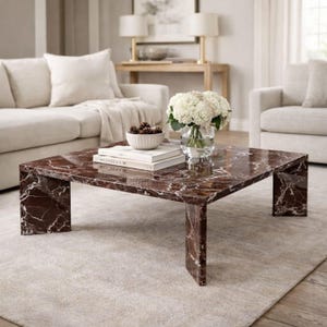 May include: A square coffee table with a rich, reddish-brown marble top and matching legs. The marble has white veining. The table is in a living room setting, with a vase of white flowers, books, and a bowl of pinecones on top.