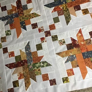 May include: A quilt featuring a repeating star pattern constructed from fabric squares in shades of orange, brown, green, and dark blue. The design incorporates white fabric strips between the blocks, creating a grid layout with autumn-themed prints.