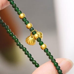 Könnte beinhalten: Ein zartes Armband mit kleinen, runden, dunkelgrünen Perlen, die mit größeren, goldfarbenen Perlen durchsetzt sind. Ein kleiner, goldfarbener Anhänger hängt an dem Armband. Das Armband wird von einer Hand gehalten.