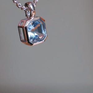 K&ouml;nnte beinhalten: Eine silberne Halskette mit einem quadratischen, hellblauen Edelsteinanh&auml;nger. Der Anh&auml;nger ist in einen silbernen Rahmen mit geometrischem Design gefasst. Die Kette ist eine filigrane Kugelkette, vor weichem Grauton.