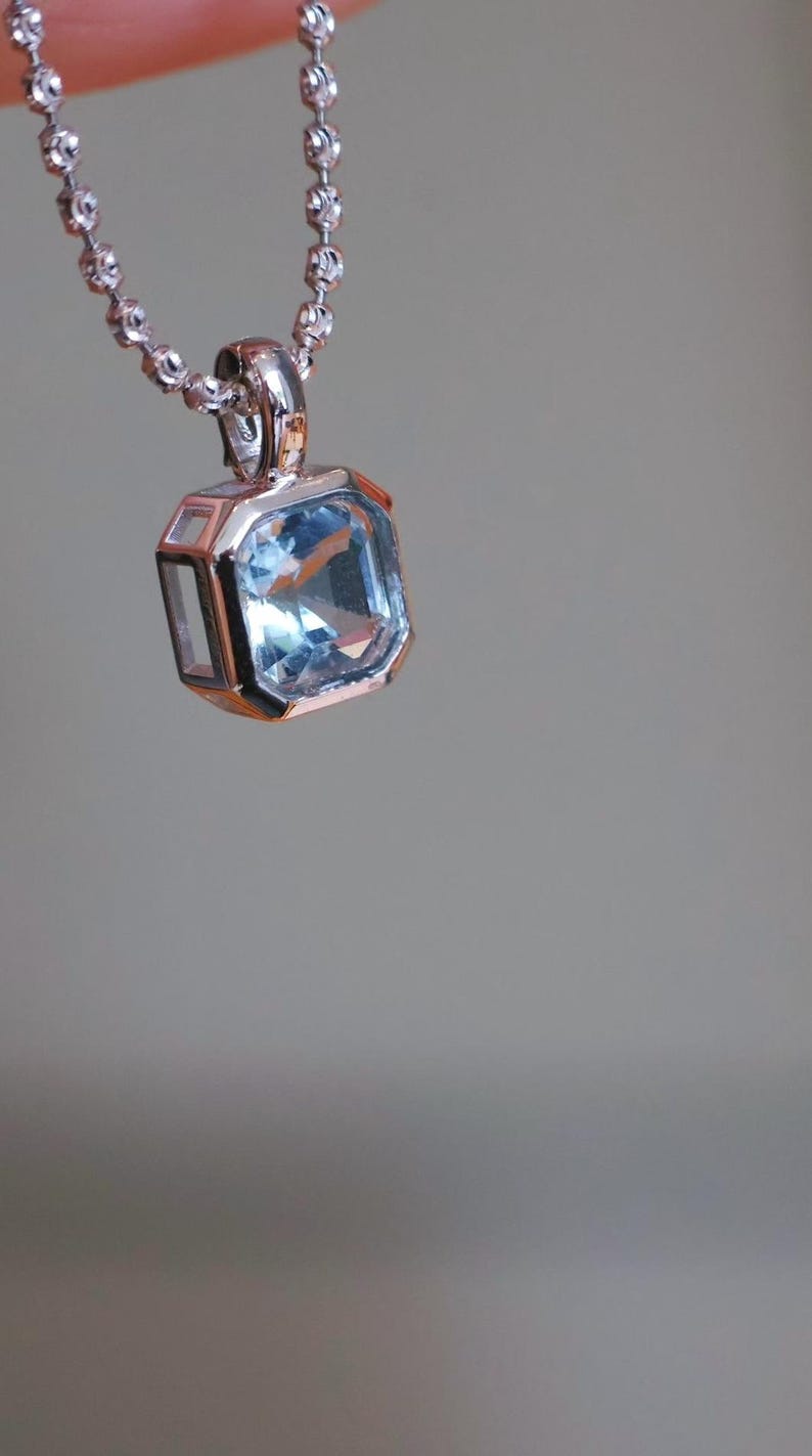 K&ouml;nnte beinhalten: Eine silberne Halskette mit einem quadratischen, hellblauen Edelsteinanh&auml;nger. Der Anh&auml;nger ist in einen silbernen Rahmen mit geometrischem Design gefasst. Die Kette ist eine filigrane Kugelkette, vor weichem Grauton.