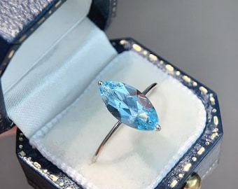 Könnte beinhalten: Ein silberner Ring mit einem hellblauen, marquiseförmigen Edelstein, präsentiert in einer marineblauen Schmuckschatulle mit weißem Satin-Innenfutter. Der Edelstein ist das zentrale Element, das das Licht einfängt.