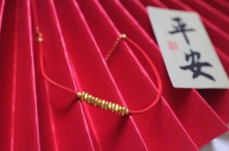 May include: A red string bracelet with gold beads. The beads are in the shape of small, flat rectangles. The bracelet is on a red background with a white card that says "平安" in black calligraphy.