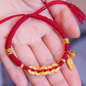 Puede incluir: Un brazalete de cuerda roja con monedas de oro y un dije. El brazalete está atado con un nudo y tiene una borla.