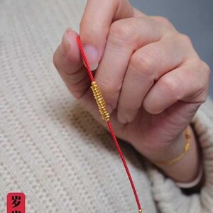 May include: A red string bracelet with a gold charm and a green bead at the end. The charm is a spiral of gold beads. The bracelet is held in a hand. The text "步步平安" is visible in the image.