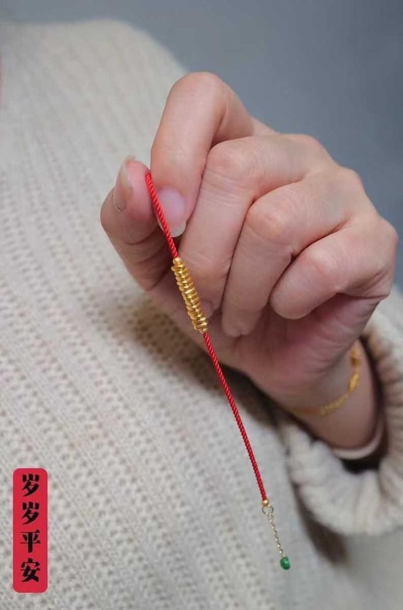 May include: A red string bracelet with a gold charm and a green bead at the end. The charm is a spiral of gold beads. The bracelet is held in a hand. The text "步步平安" is visible in the image.
