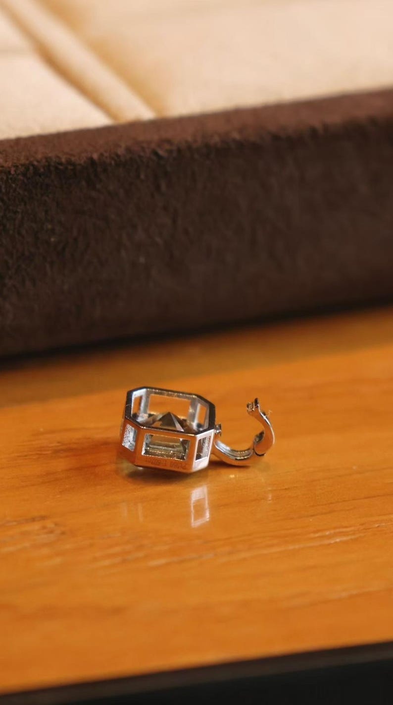 K&ouml;nnte beinhalten: Ein silberner Ohrring mit geometrischem Design, der eine rechteckige Form mit einem kleinen, klaren Edelstein aufweist. Der Ohrring ist offen und zeigt den Verschluss. Der Ohrring liegt auf einer Holzoberfl&auml;che.