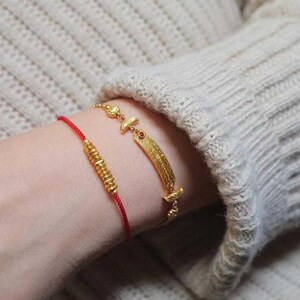 May include: A red string bracelet with a gold charm bracelet featuring a small pendant and a larger, flat, rectangular pendant. The bracelet is worn on a person's wrist.
