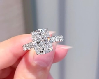 Könnte beinhalten: Ein funkelnder Verlobungsring mit einem großen, rechteckig geschliffenen Diamanten. Der Ring hat ein Band aus Platin oder Weißgold, mit kleineren Diamanten entlang des Bandes und auf beiden Seiten des Hauptsteins. Der Ring wird von einer Hand gehalten.