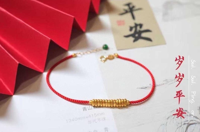 May include: A red string bracelet with a gold charm that says "Sui. Sul. Ping, an" in Chinese characters. The charm is made of a series of stacked gold rings. The bracelet is on a white surface with a red folded paper background.