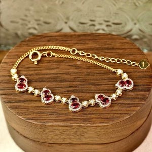 May include: A gold bracelet with a chain link design and red and white gemstones. The bracelet is displayed in a wooden box.