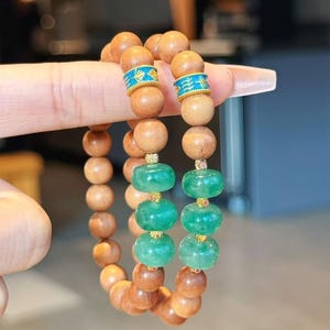 May include: Two bracelets with wooden beads and green jade beads. The wooden beads have gold accents with blue and gold designs.