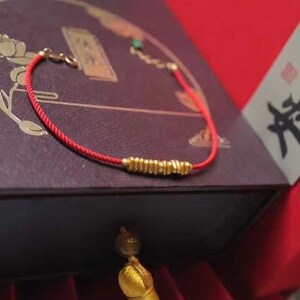 May include: A red string bracelet with a gold charm. The bracelet is on a dark brown box with Chinese characters. The box is labeled "Sui. Sui. Ping. An."